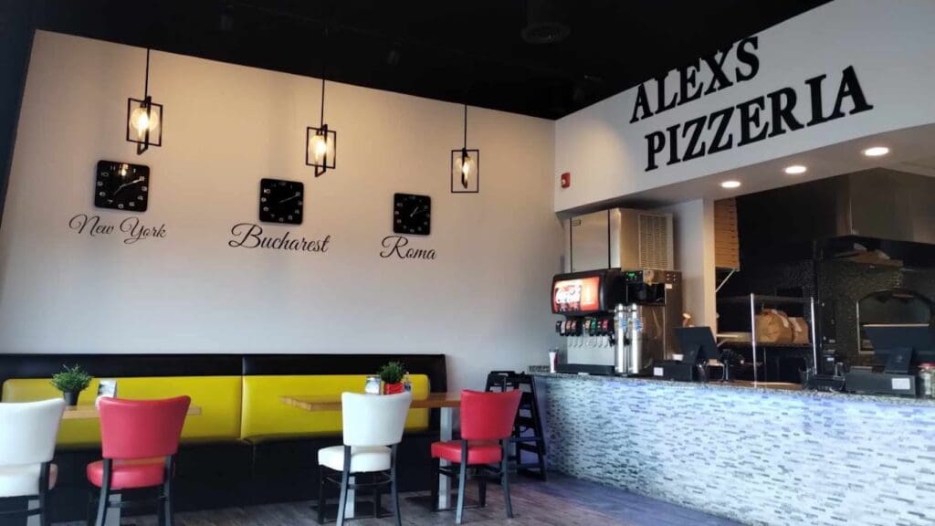 A modern pizzeria interior features contemporary seating with colorful accents, pendant lighting, and a service counter in a minimalist aesthetic.