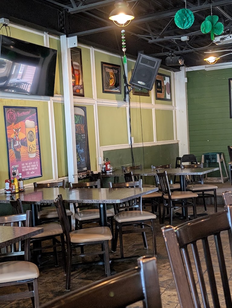 An empty restaurant dining room with sage green walls, wooden tables and chairs, framed artwork, and ambient ceiling lighting.
