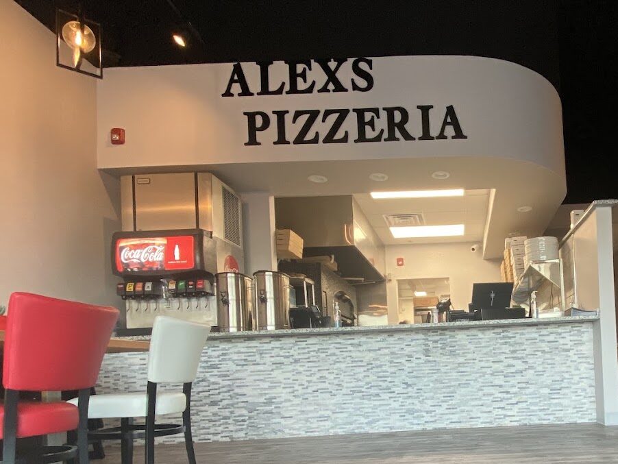 An empty pizzeria interior at night featuring a service counter, seating area with red chairs, and warm lighting throughout the modern space.