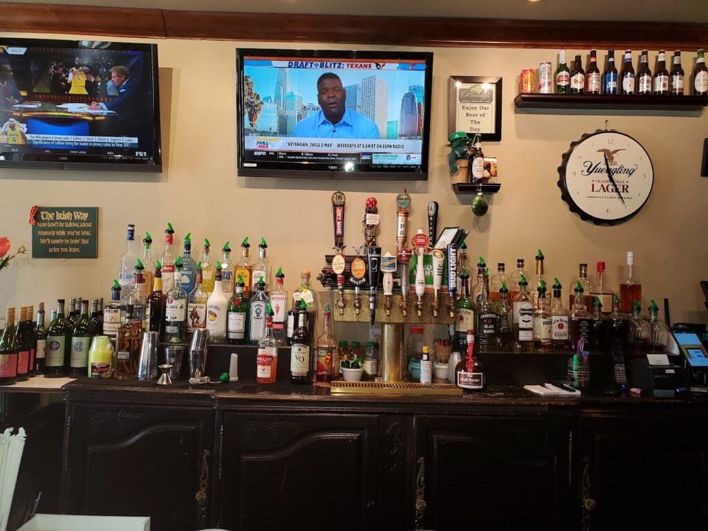 A well-stocked bar displaying numerous liquor bottles on wooden shelves with a television and decorative clock on the wall above.
