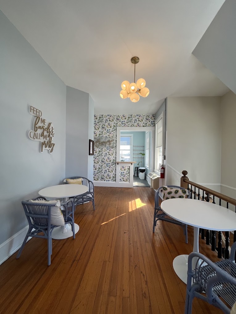 A bright, minimalist nursery room features a white crib, wooden desk, and pendant light fixture along a hallway with warm hardwood flooring and neutral wall tones.