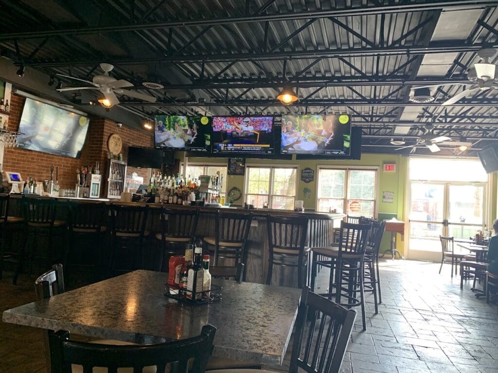 An empty bar or restaurant interior with wooden counters, black chairs, industrial ceiling, and natural light streaming through windows.