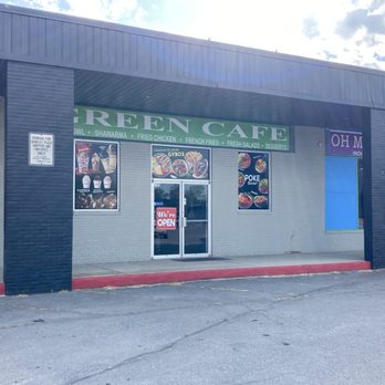 A small Green Cafe storefront with signage and window displays located in a commercial strip mall during daytime.