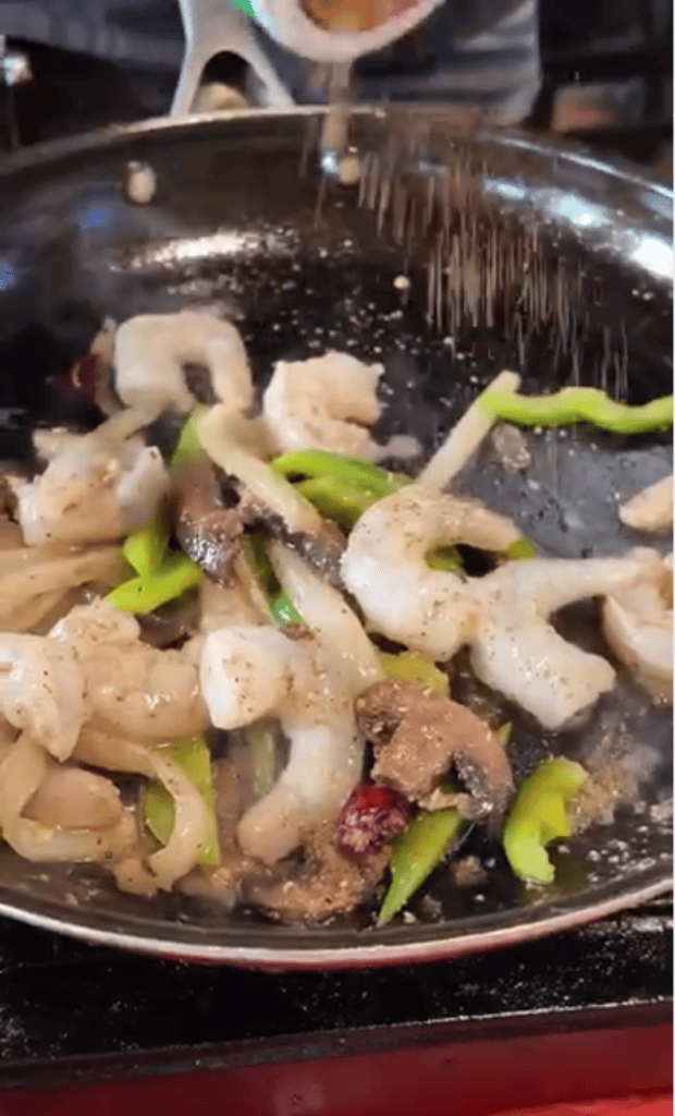 A chef stir-fries shrimp and vegetables in a wok while pouring sauce, captured in an action close-up shot.