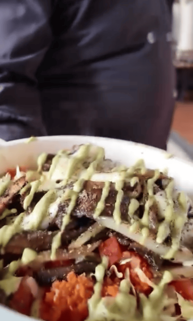 A close-up view of a plated taco or sandwich topped with shredded cheese, lettuce, and tomatoes with a white sauce drizzle, with a person partially visible in the background.