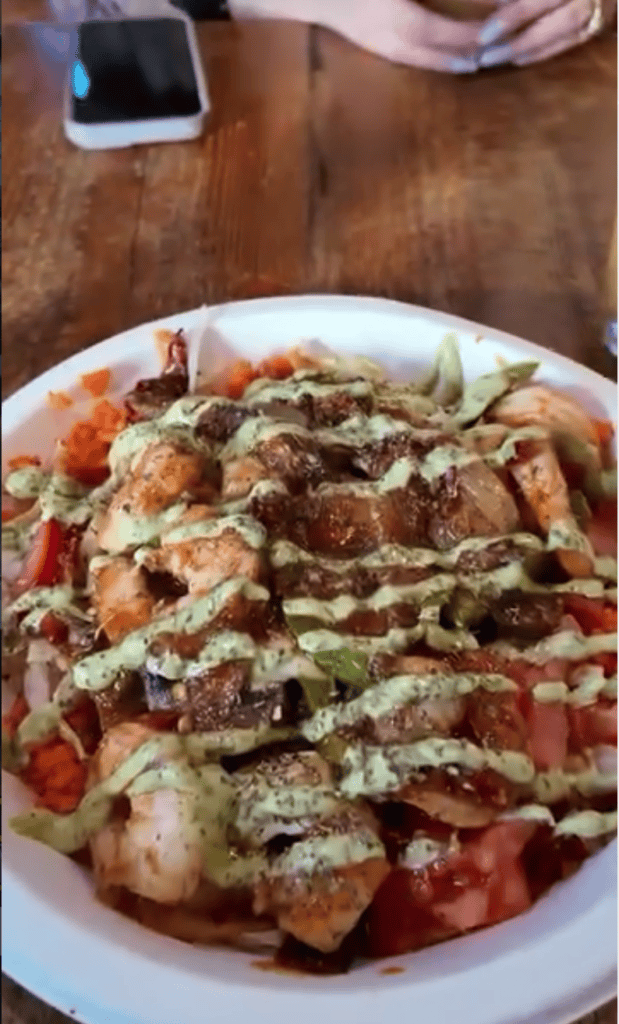 A close-up overhead view of a freshly prepared pizza with toppings on a white plate, placed on a wooden table with a smartphone visible in the background.