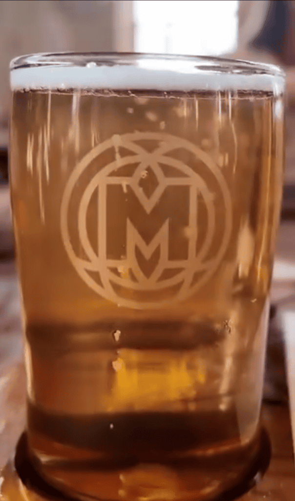 A close-up photograph of an amber-colored beverage in a glass with an engraved circular logo or crest.