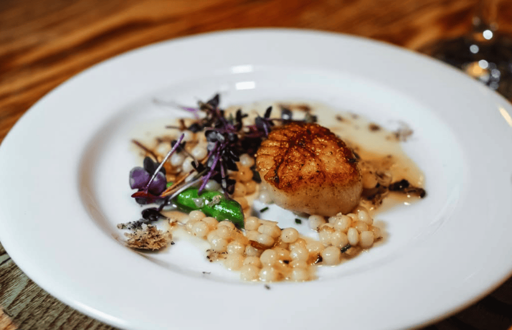 An elegantly plated fine dining dish featuring a seared protein with grain accompaniment and fresh microgreens on a white plate against a warm wooden surface.