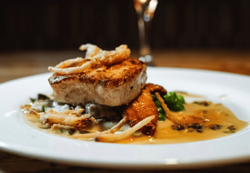 A beautifully plated seared steak with golden crust sits atop a savory sauce with vegetables and garnish on a white plate in an upscale restaurant setting.