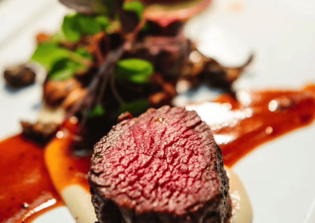 A close-up of a perfectly seared beef steak garnished with microgreens, artfully plated with sauce on a white dish.