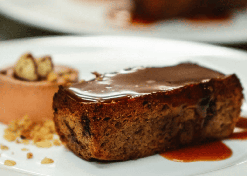 A close-up photograph of an elegantly plated chocolate cake with glossy ganache topping and caramel sauce on a white plate.