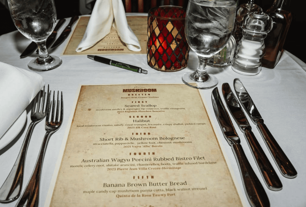 An overhead view of an elegant formal dinner place setting with a vintage menu card, fine dining silverware, and crystal glassware arranged on a white tablecloth.
