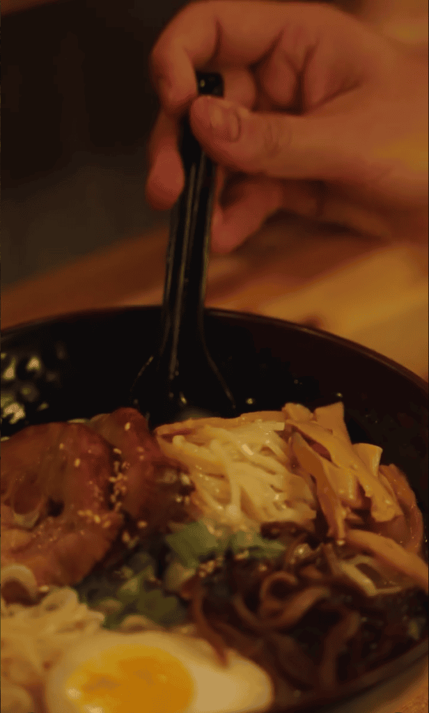 A close-up shot of someone eating noodles from a dark bowl using chopsticks in warm indoor lighting.