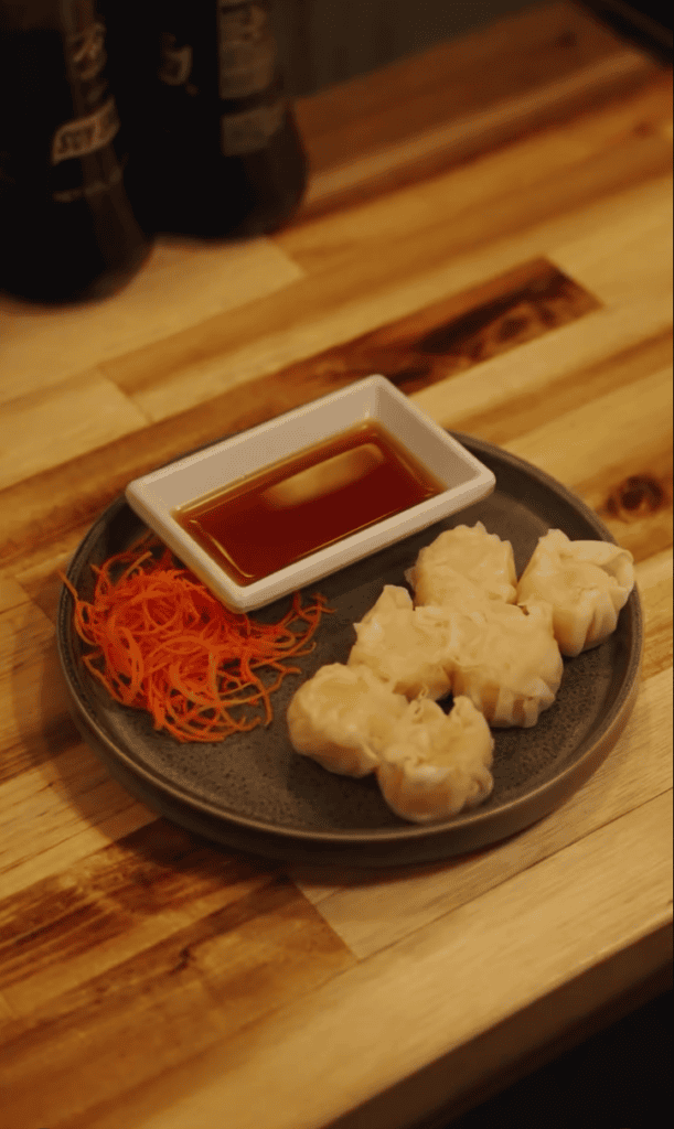 A overhead view of Asian fried appetizers with red dipping sauce arranged on a dark ceramic plate on a wooden table with beer bottles in the background.