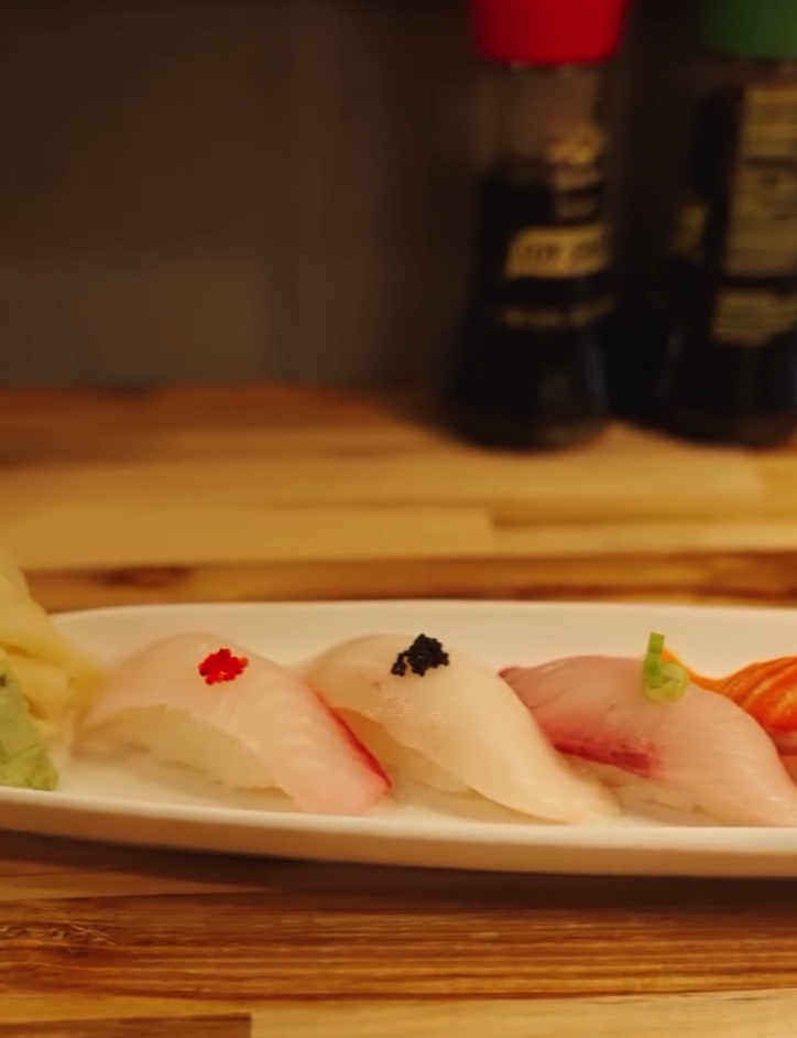 A carefully arranged sashimi platter with fresh sliced fish is displayed on a white plate on a wooden surface with wine bottles visible in the background.
