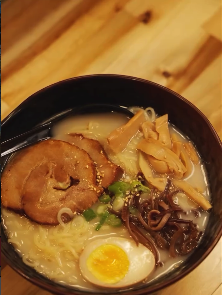 A steaming bowl of ramen with noodles, broth, sliced pork, and a soft-boiled egg sits in a black bowl on a wooden surface.