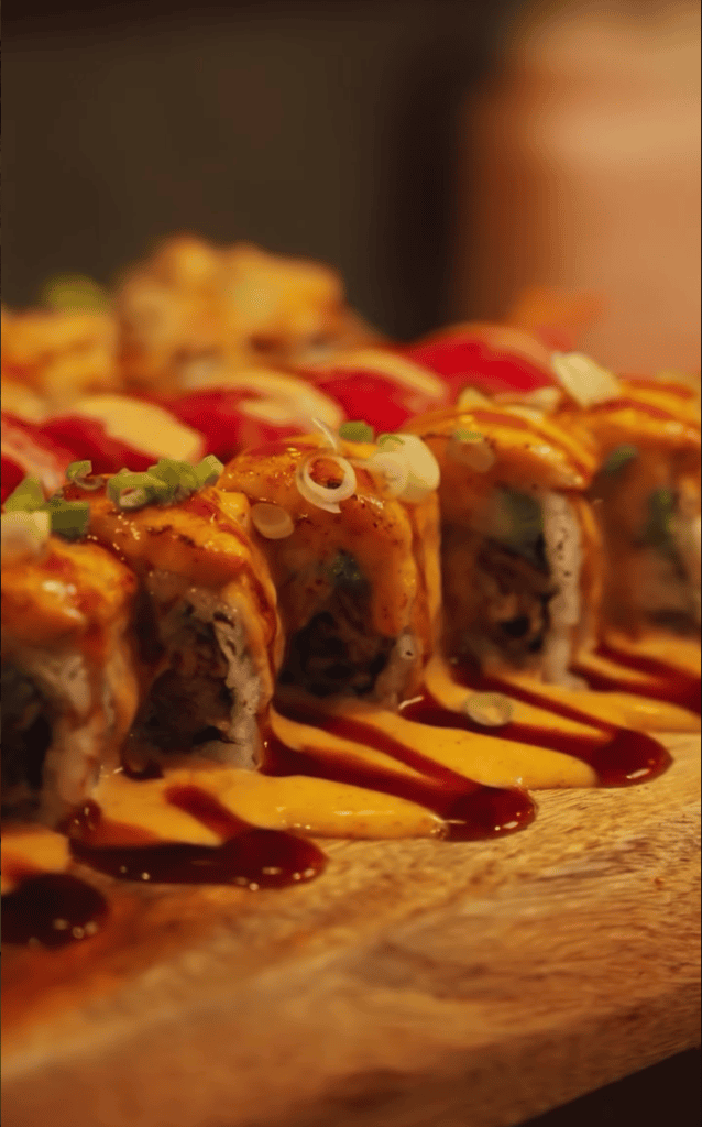 A beautifully plated row of sushi rolls on a wooden board with artistic red sauce drizzle, photographed in warm golden lighting.