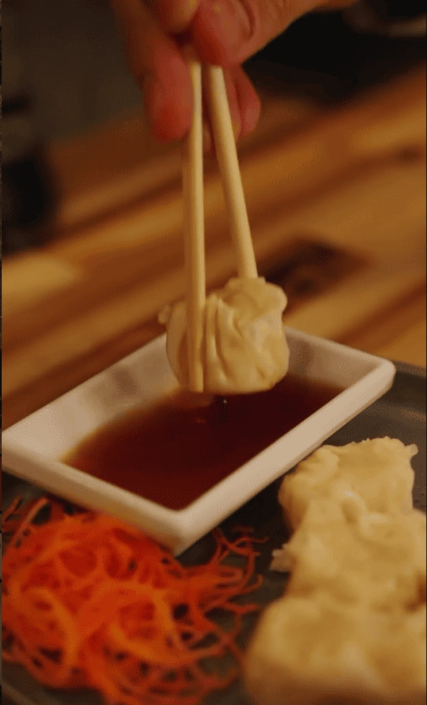 A close-up shot of Asian dumplings being lifted with chopsticks over a small bowl of dipping sauce, with fresh ginger visible on the side.