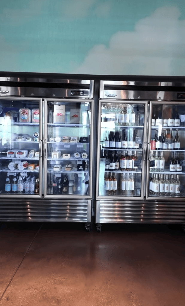 A commercial double-door refrigerator display case filled with beverages and products, photographed against a blue-tinted sky background in what appears to be an indoor commercial kitchen or storage facility.