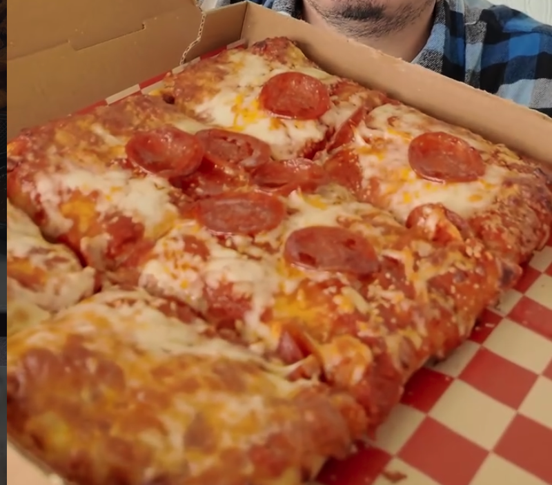 A person displays a freshly made pepperoni pizza in an open box lined with red and white checkered paper.