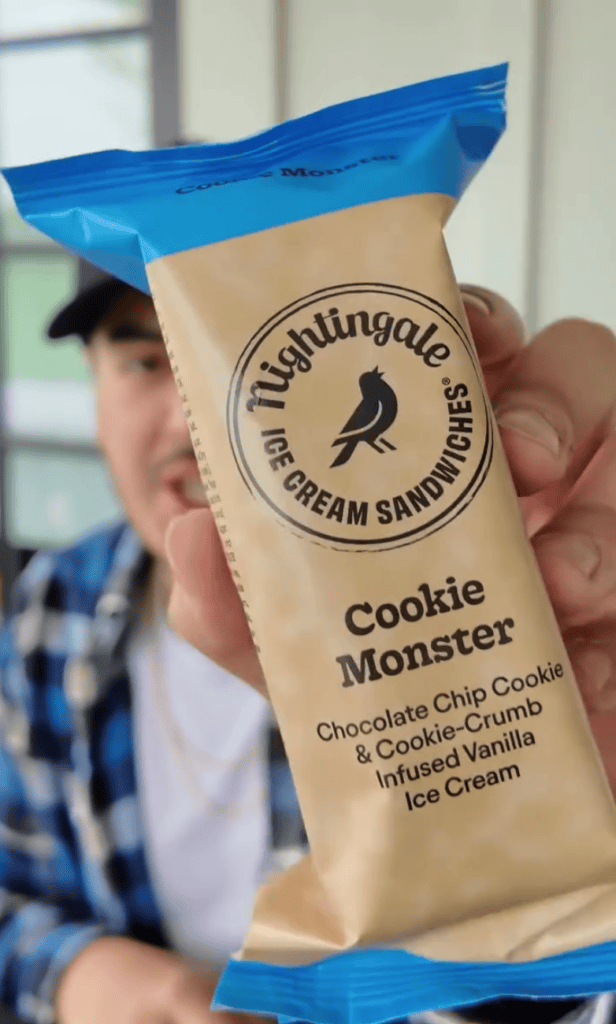 A close-up of a hand holding a packaged Cookie Monster sandwich cookie with blue packaging and a visible product label.