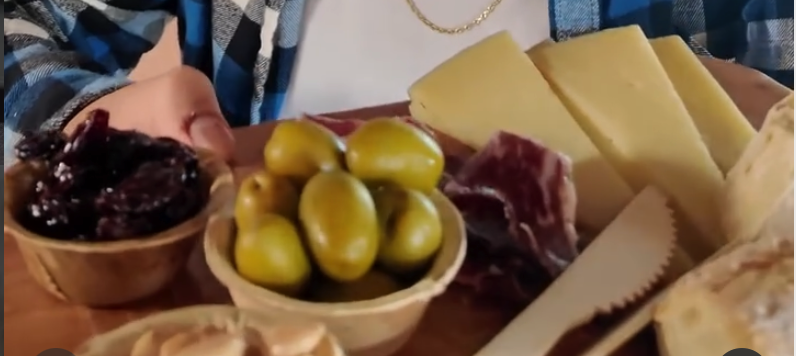 An overhead view of a rustic charcuterie board featuring cured meats, cheese, pickles, and olives arranged on a wooden surface with someone's hands visible in the frame.