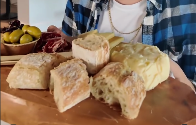 A person in a blue plaid shirt displays freshly baked bread rolls on a wooden table, with olives and cured meats visible in the background.