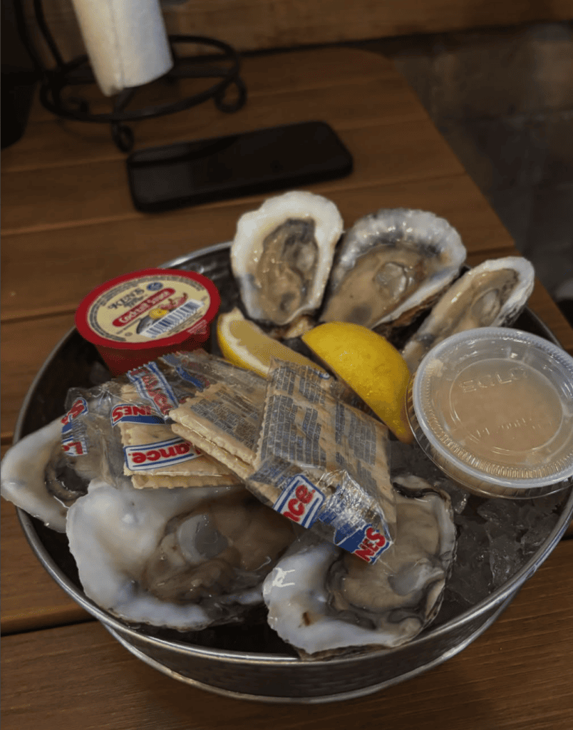 An overhead view of a seafood platter featuring fresh oysters, bread, and various condiments arranged on a round white plate on a wooden table.
