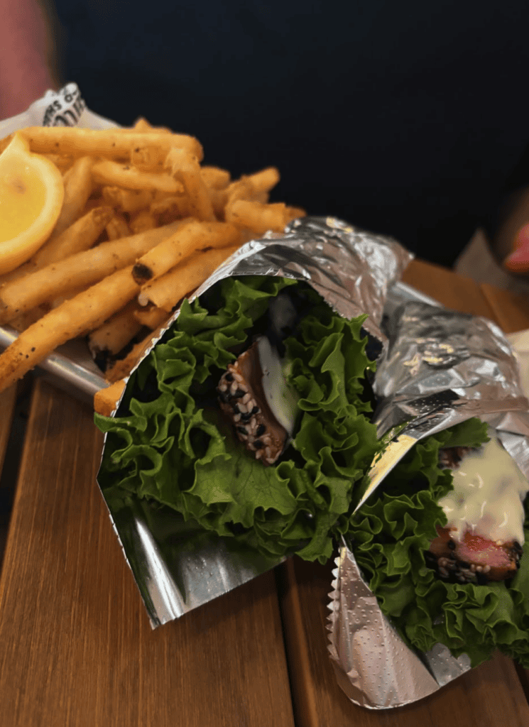 A foil-wrapped sandwich filled with greens and meat is served alongside crispy golden fries and a lemon wedge on a wooden surface.