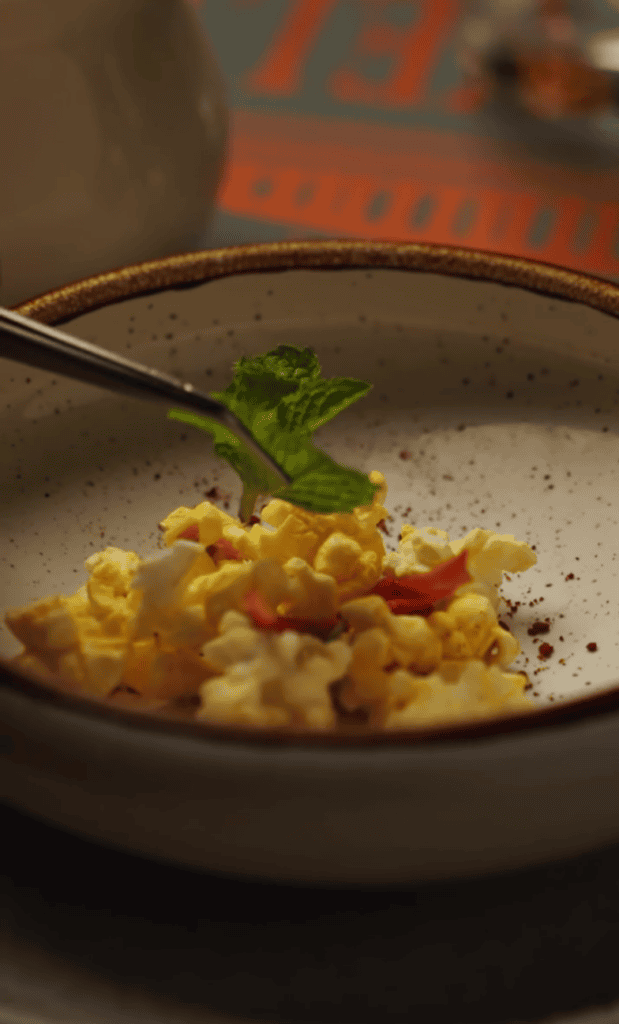 A close-up shot of freshly cooked scrambled eggs in a pan, garnished with green herbs and black pepper, captured during food preparation.