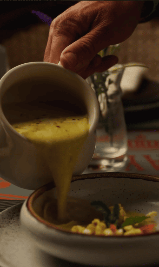 A person pours a golden sauce or gravy from a bowl onto a plated dish in an indoor dining setting.