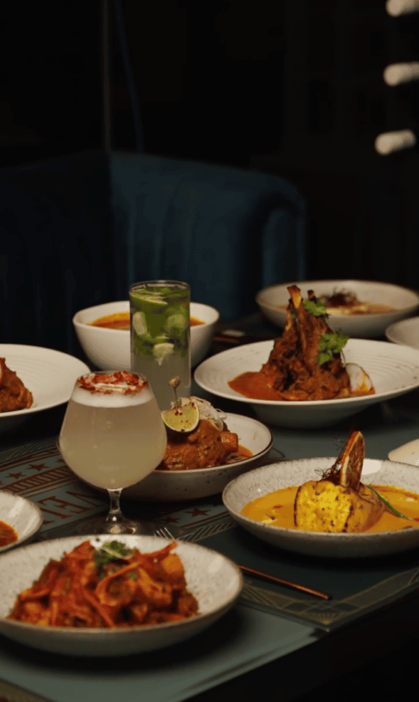 An elegant fine dining spread featuring multiple plated dishes with fresh vegetables, a cocktail, and sophisticated tableware in a dimly lit restaurant setting.
