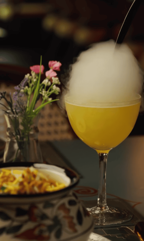 A close-up of a golden cocktail in an elegant glass beside fresh pink flowers and a plated dish in a dimly lit dining setting.