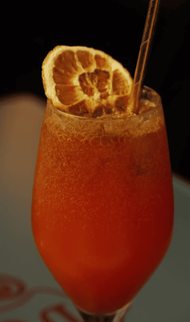 A vibrant coral-orange cocktail garnished with a dried citrus wheel and served with a straw against a dark background.