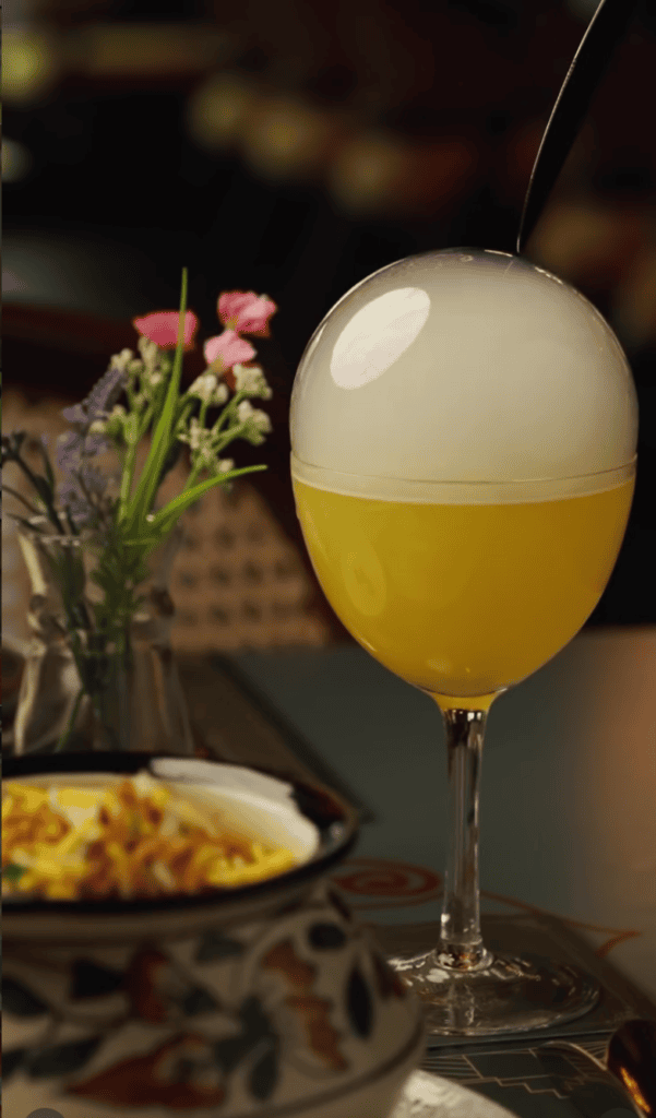 A golden-yellow cocktail in a tall glass sits elegantly on a table beside pink flowers and a plated dish in warm indoor lighting.