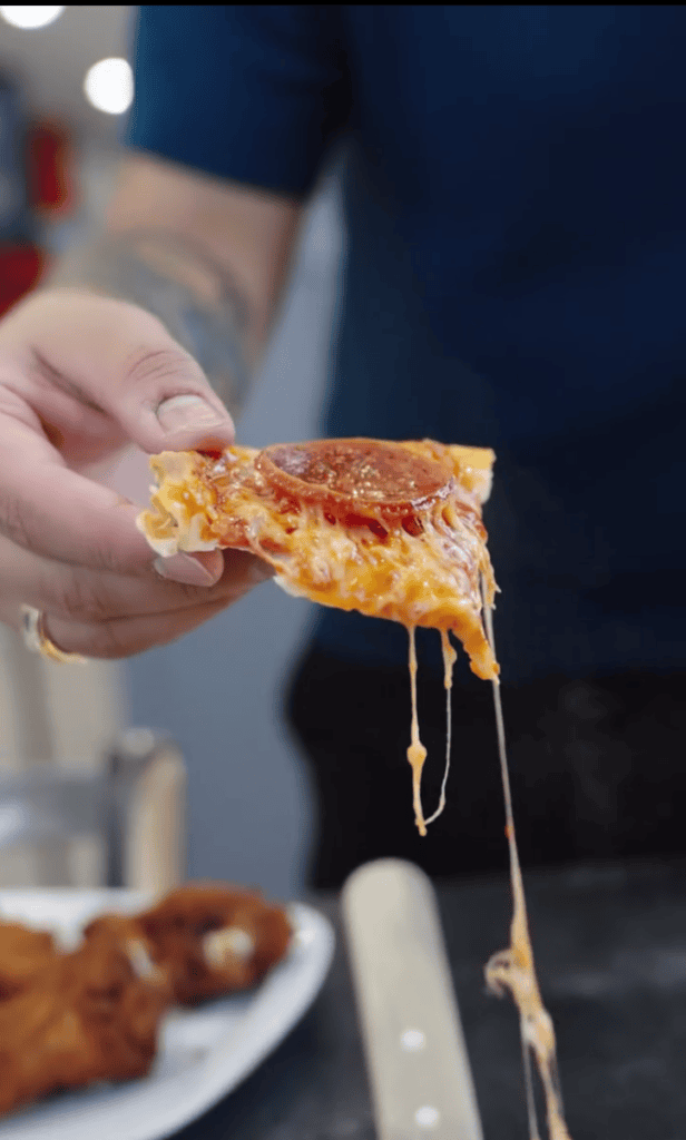 A person holds a slice of hot pizza with melted cheese stretching from the bite, captured in a close-up shot.