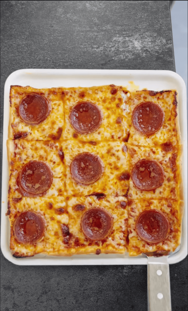 A freshly baked pepperoni pizza in a white rectangular baking dish sits on a dark gray countertop, cut into nine squares.
