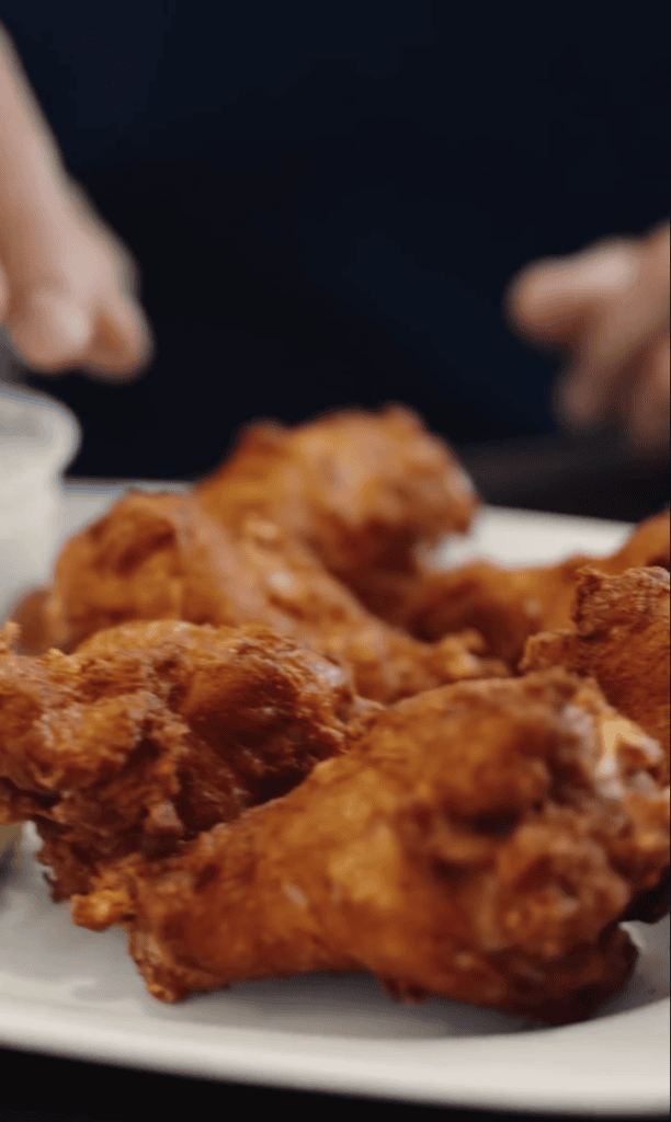 Golden-brown fried chicken pieces are arranged on a white plate with hands visible at the edges against a dark background.