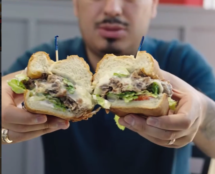 A person displays two large, generously-filled sandwiches with lettuce and other ingredients in a close-up food presentation shot.