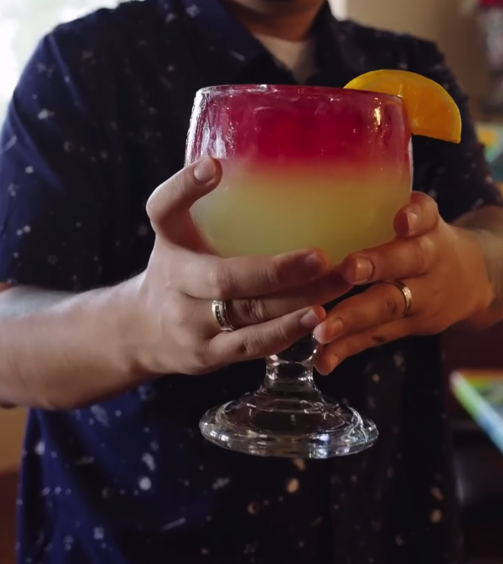 A person holds an elegant layered cocktail with red and pale yellow layers, garnished with a citrus wheel, against a dark blue outfit.