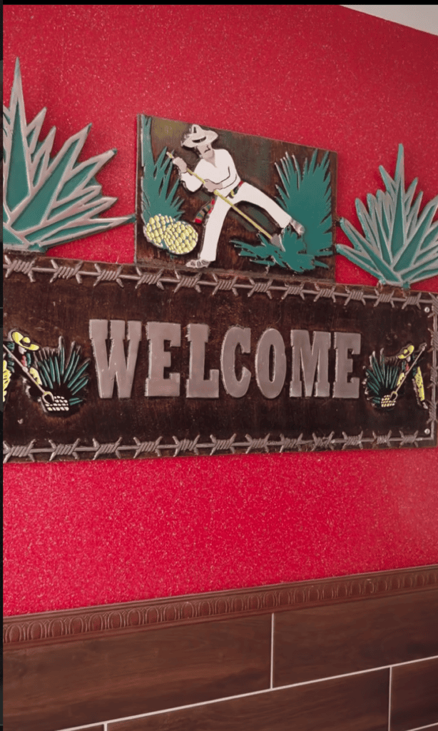 A Western-themed welcome sign featuring a cowboy figure with agave plants and barbed wire decoration against a red and brown background.