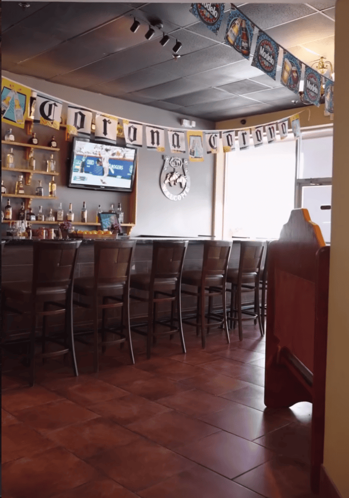 An empty bar interior featuring dark wooden bar stools, shelving with bottles and memorabilia, decorative bunting overhead, and warm wooden tones throughout.