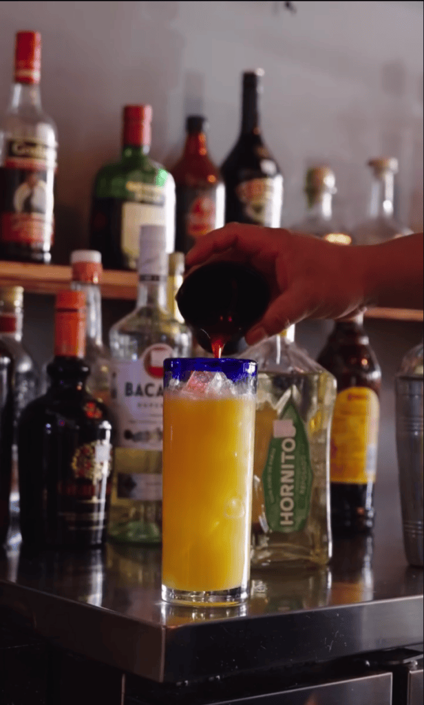A bar counter displays colorful cocktails and spirits with a red bartender's hat perched on a glass, suggesting an upscale drinking establishment.