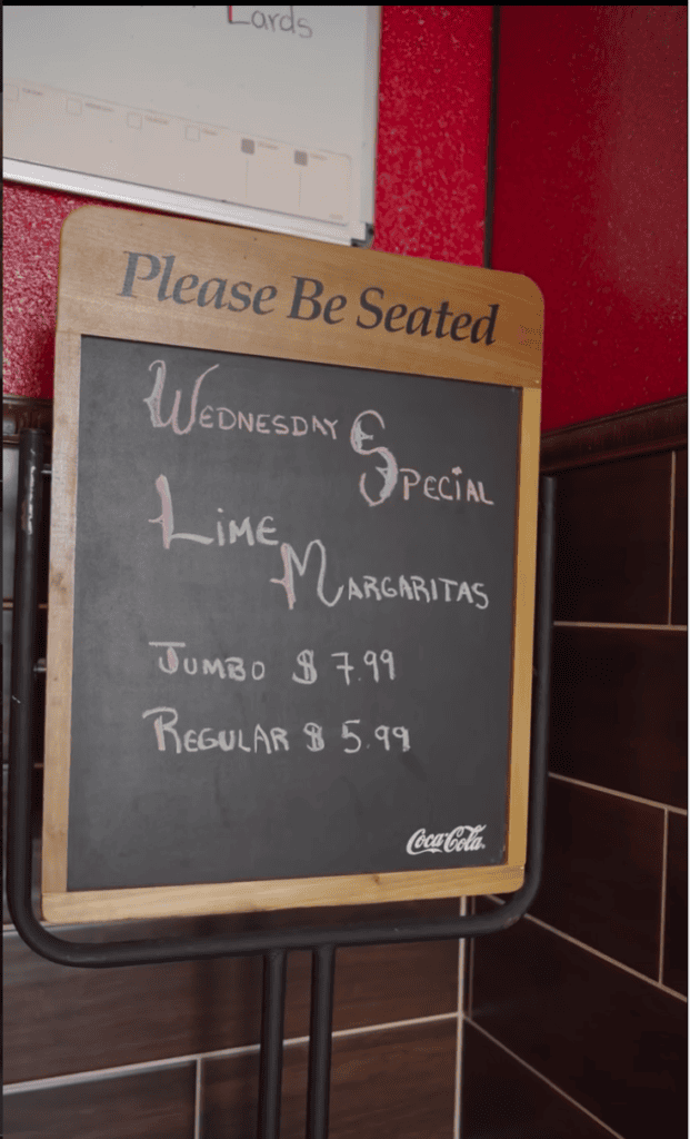 A wooden-framed chalkboard sign with handwritten text stands at a venue entrance against red fabric, displaying seating information and event details.