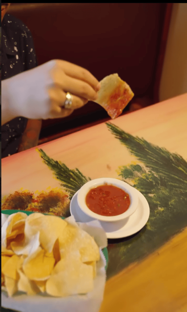 A person's hand dips a crispy chip or bread into a red sauce at a dining table with asparagus and other appetizers.