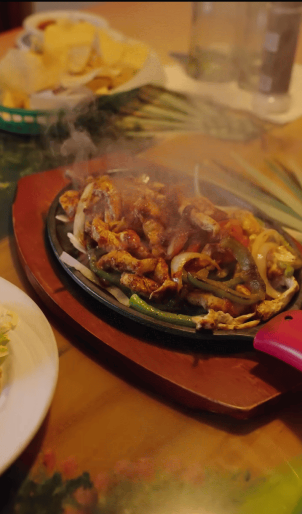 A sizzling plate of fajitas served on a hot skillet at a dining table with accompaniments and beverages visible.