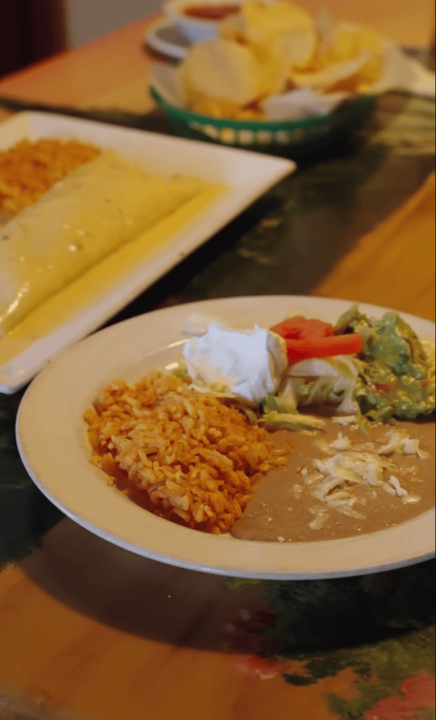 A restaurant table displays a plate of food featuring rice, vegetables, and soup alongside bread and other dishes in warm indoor lighting.