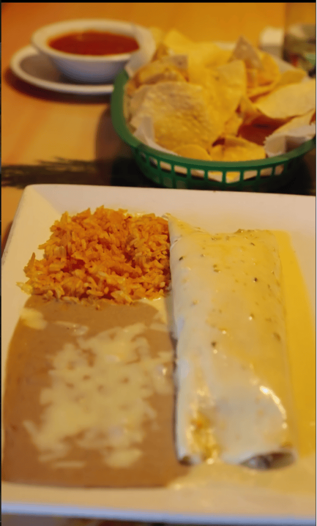 A plate of Mexican-style food featuring enchiladas, rice, and cheese sauce, served alongside chips and salsa in a restaurant setting.