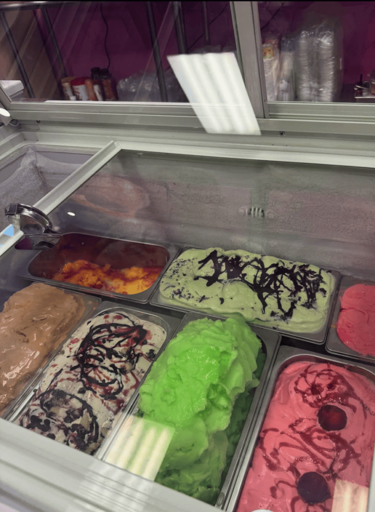 A vibrant ice cream shop display case showing multiple colorful frozen dessert flavors in bright greens, pinks, browns, and purples.