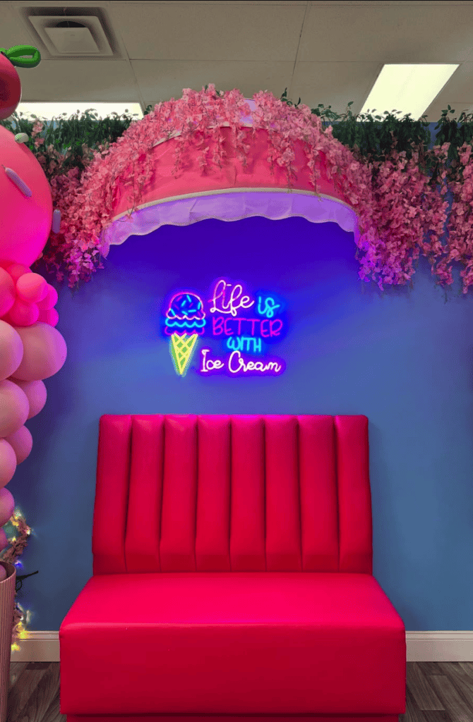 A vibrant party photo booth setup featuring a red velvet sofa beneath pink floral arrangements and a glowing neon sign against a blue wall.